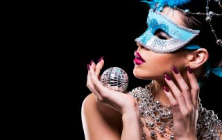 beautiful woman with dark makeup wearing blue and silver mask surrounded by party accessories on light black background