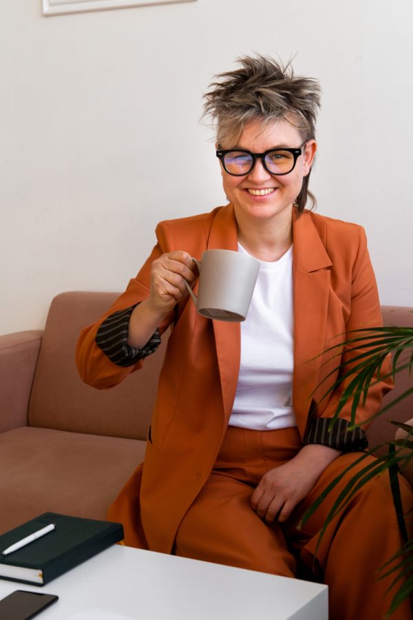young non binary homosexual person wearing orange color suit sitting on sofa, holding cup of coffee