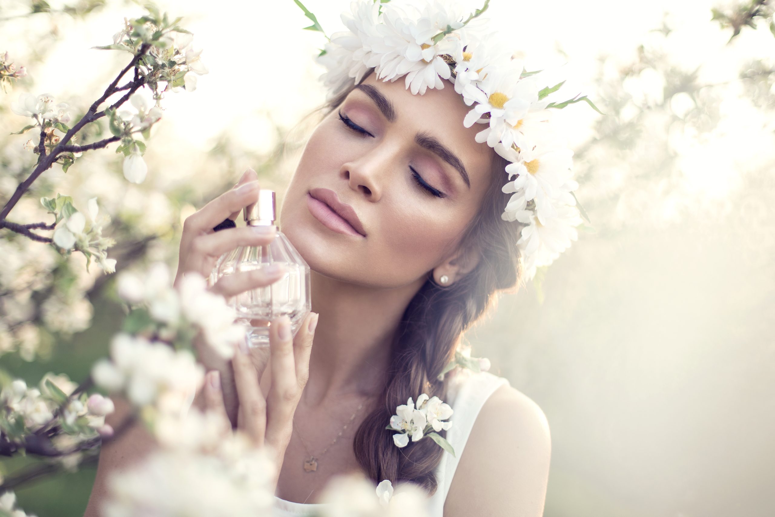 Beautiful sensual woman in a white flower crown standing next to a blooming tree with white flowers and smelling a bottle of perfume
