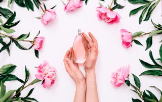 top view of pink bottle of women's perfume in female hands and peony flowers around on white background.